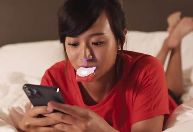 Cô gái đang sử dụng bộ dụng cụ làm trắng răng khi đang sử dụng điện thoại trên giường